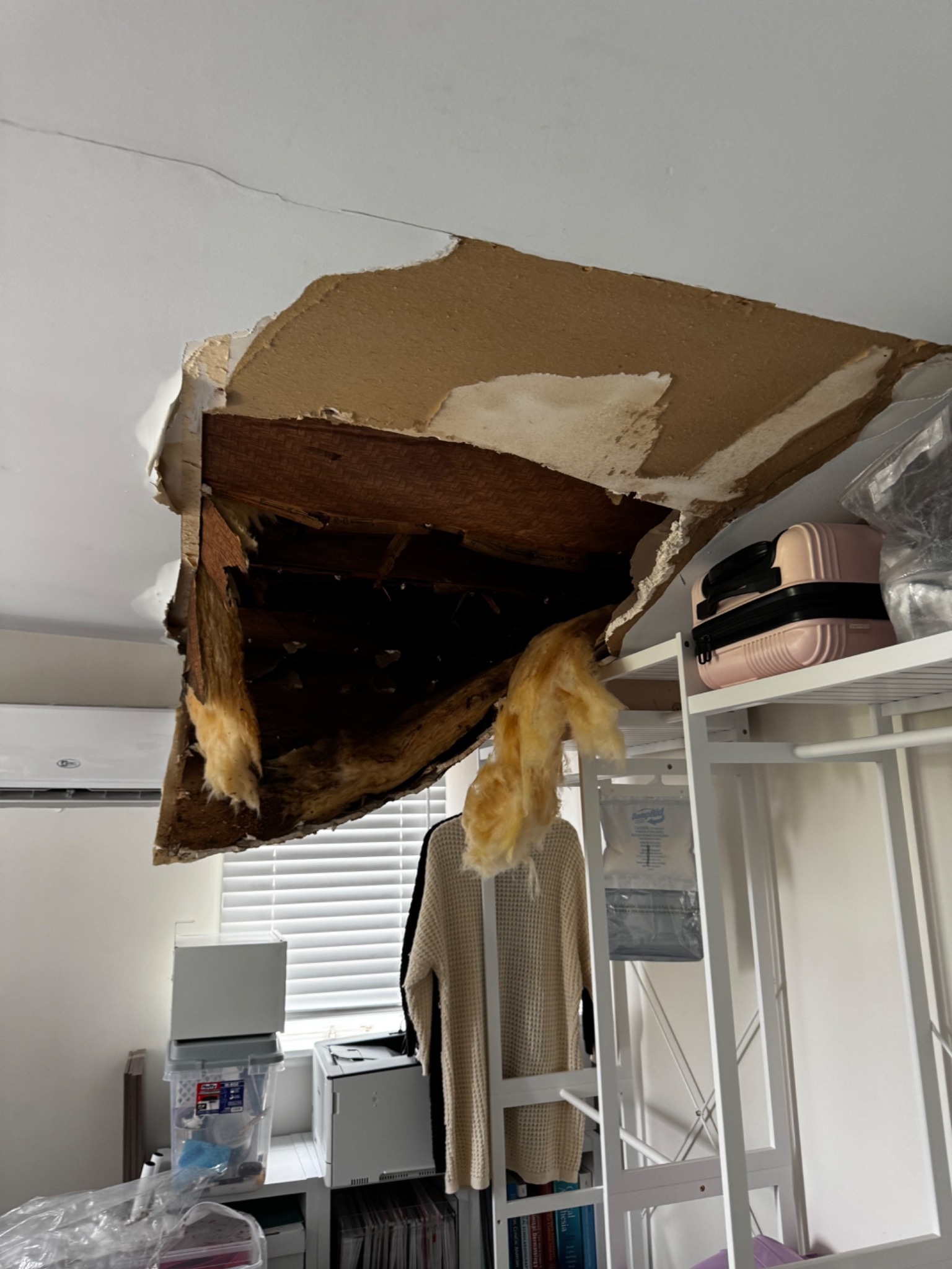 A large section of ceiling and roof collapsed directly above a bed, exposing dark framing and hanging insulation.